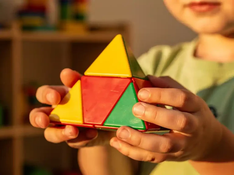 Kind houdt kleurrijke pyraminx-puzzel vast met gele, rode en groene vlakken, verlicht door warm middaglicht in speelkamer.