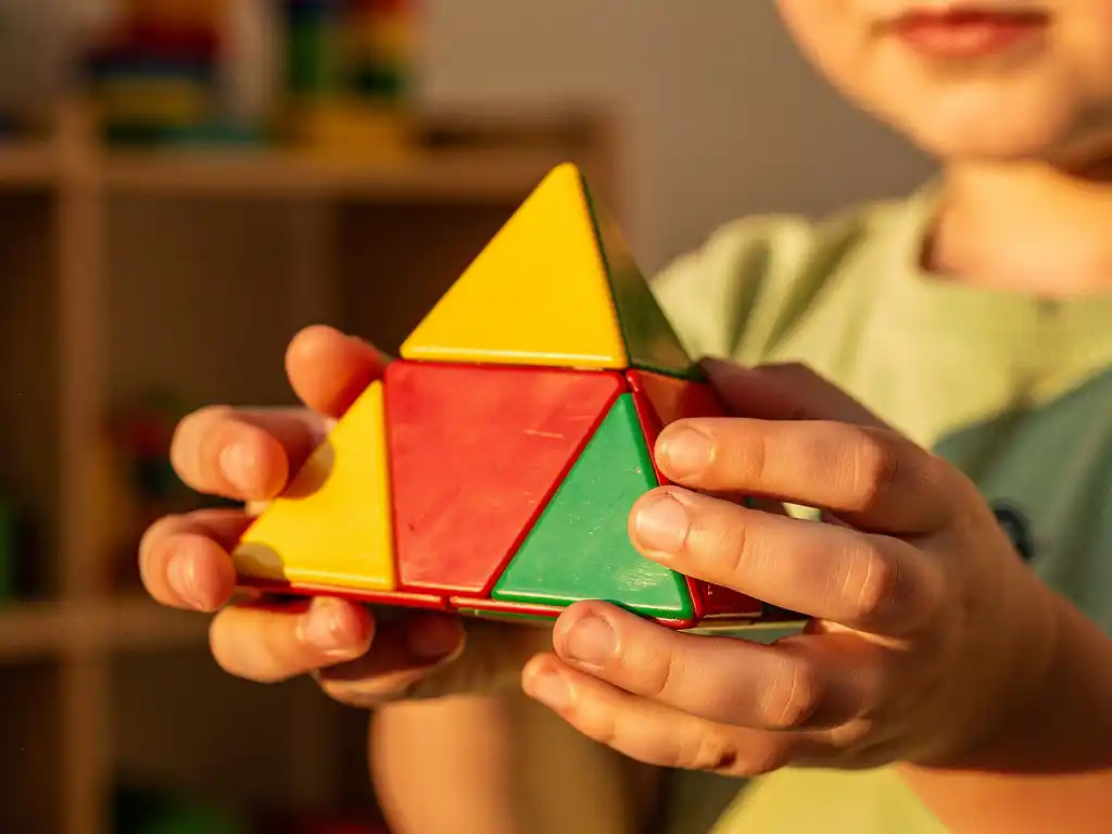 Kind houdt kleurrijke pyraminx-puzzel vast met gele, rode en groene vlakken, verlicht door warm middaglicht in speelkamer.