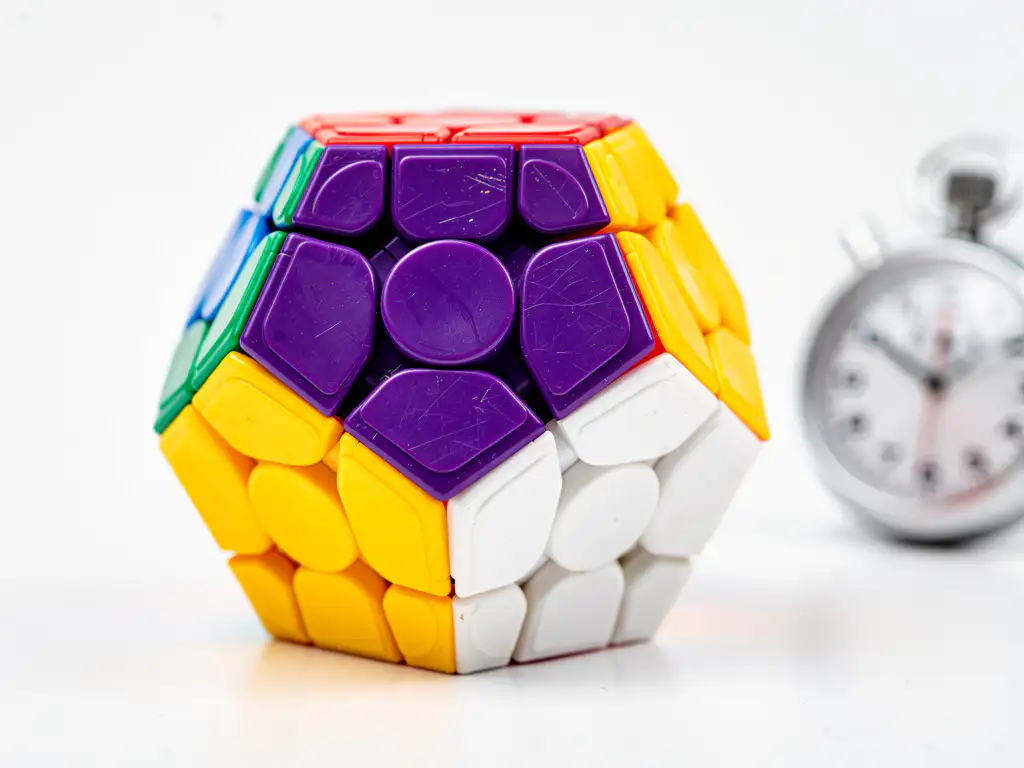 Kleurrijke Megaminx dodecaëder puzzel op wit oppervlak met onscherpe stopwatch, paarse en gele pentagonale tegels in close-up.