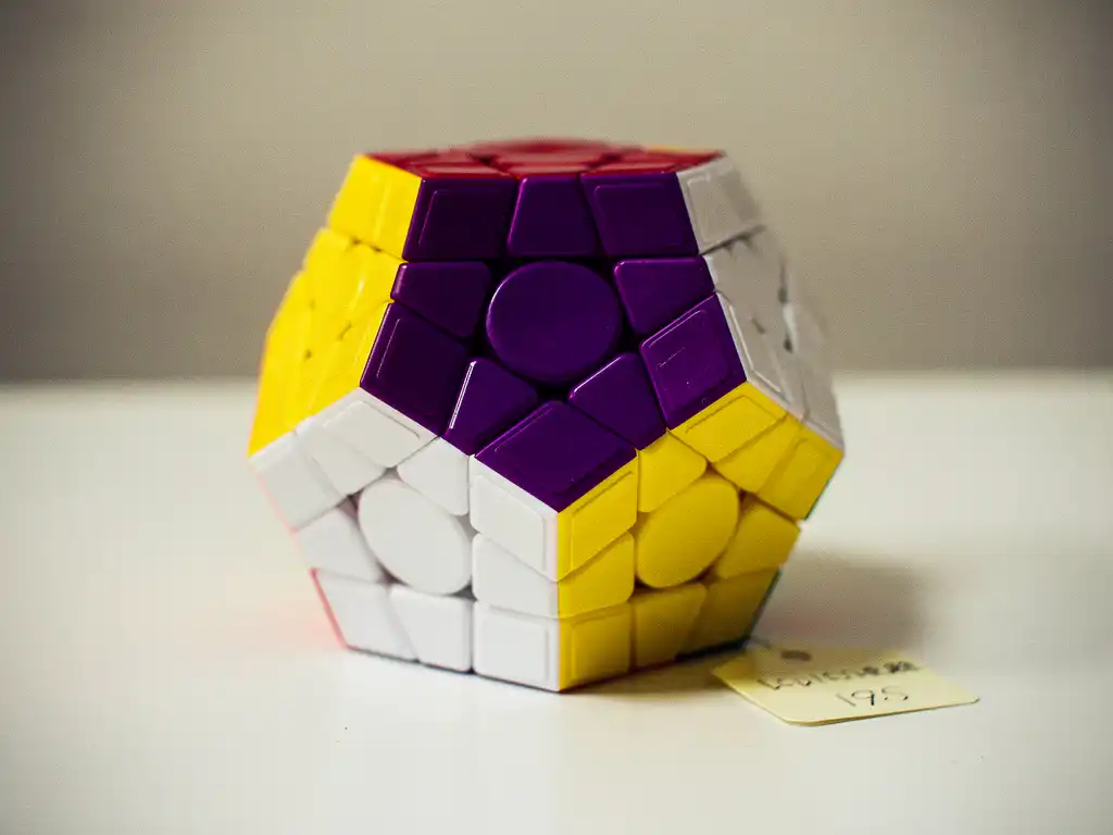 Opgeloste megaminx puzzel met twaalf pentagonale vlakken in paars, geel en wit op een wit bureau.