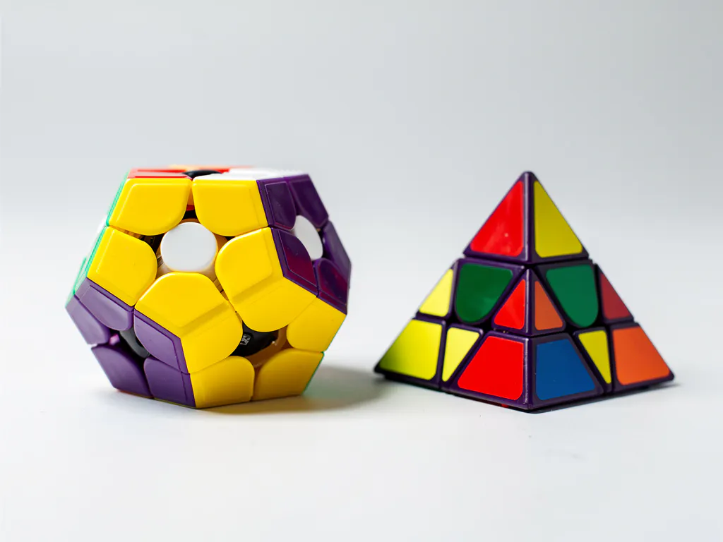 Glanzende megaminx en kleurrijke pyraminx naast elkaar op wit oppervlak, verlicht met zacht studiolicht.