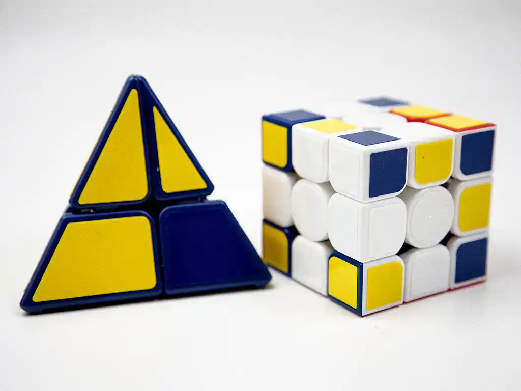 Pyraminx en skewb puzzels naast elkaar op wit oppervlak, met gele, blauwe en witte stickers in studioverlichting.